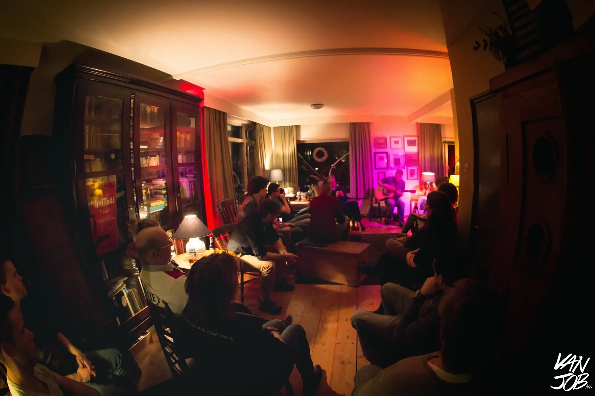 Huiskamerconcert vanuit het publiek — warme sfeerverlichting, iedereen luistert aandachtig
